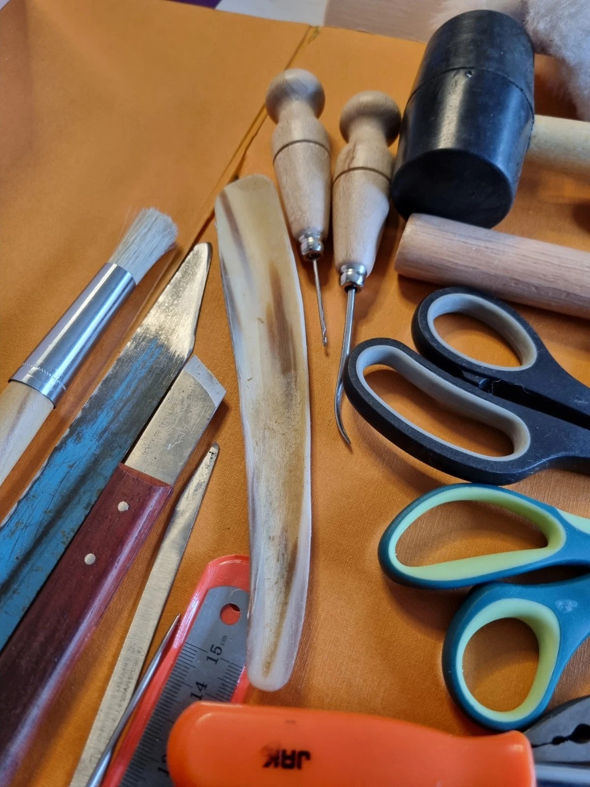 A collection of leatherworking and crafting tools laid out on a tan leather surface, including a pair of black and blue-handled scissors, a wooden mallet, a black cylindrical hammer, a bone chisel, a metal file, a small knife with a reddish-brown handle, and other hand tools.