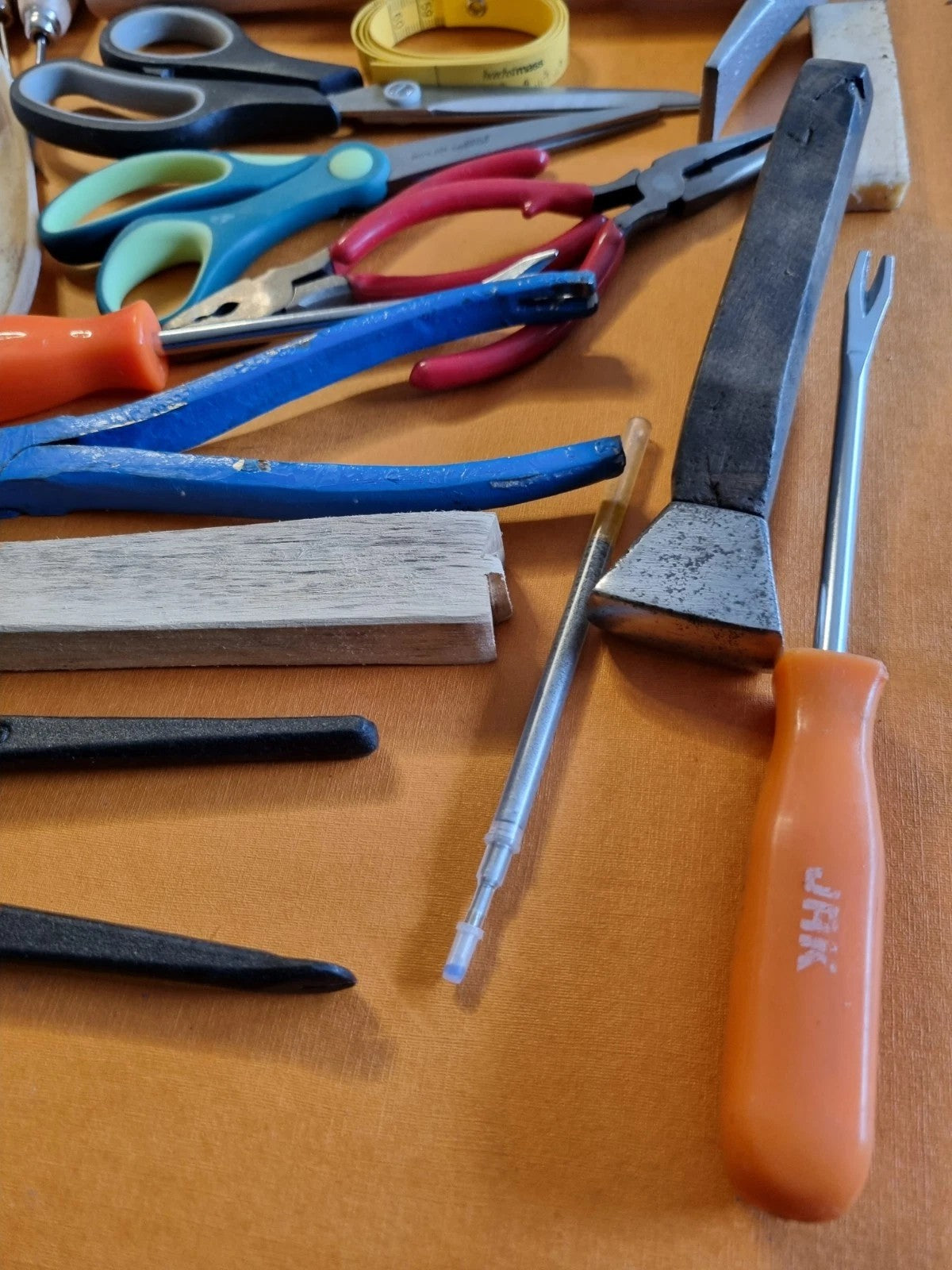 A collection of hand tools with various colored handles, including orange, blue, pink, and black, laid out on a brown surface. Visible brands include JAK on the orange-handled tool and Dremel on the white text on the metal rod.