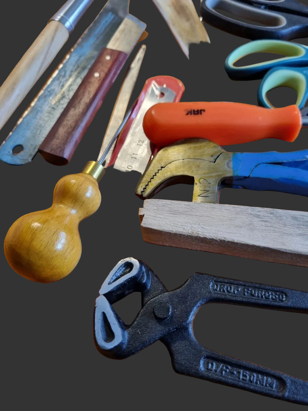 A collection of hand tools and measuring instruments arranged on a wooden surface, including a wooden gourd-handled ruler, a metal ruler, a wooden square, a black clamp tool, a blue-handled tool, an orange-handled tool, and scissors with blue and green handles.