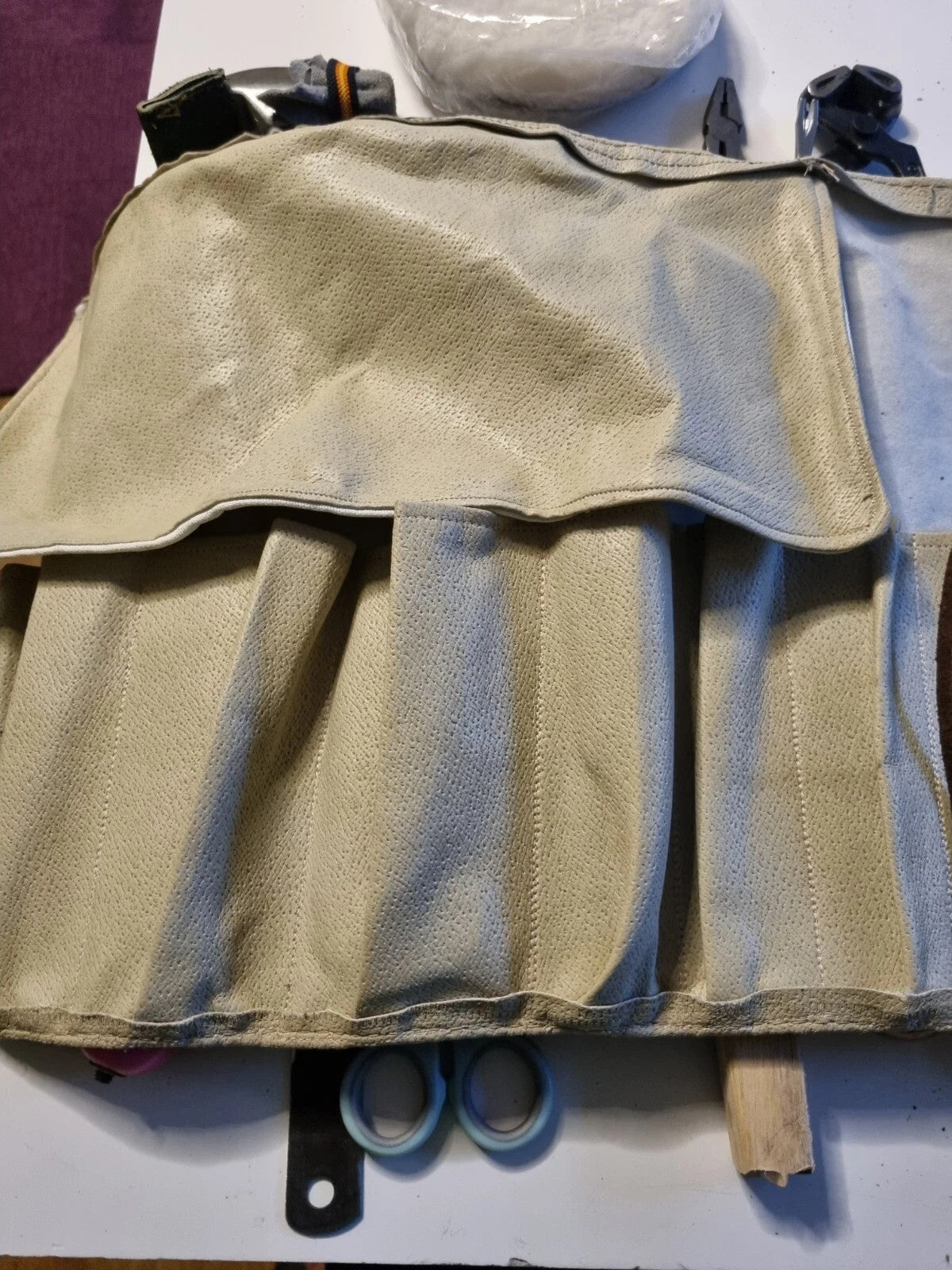 A beige canvas tool roll or organizer pouch laid flat on a white surface, with several small tools partially tucked into or around it, including blue-handled scissors and a wooden stick.