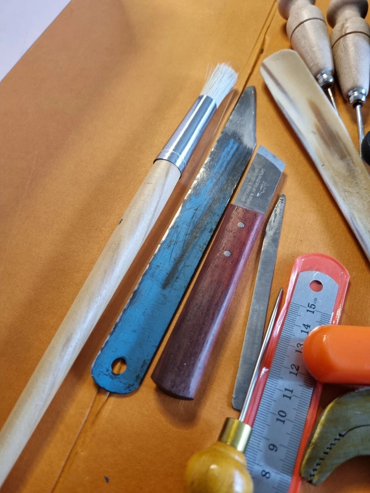 A collection of leatherworking and carving tools laid out on a tan leather surface, including a brush, knives, a bone chisel, a rasp, and a small orange-handled tool.