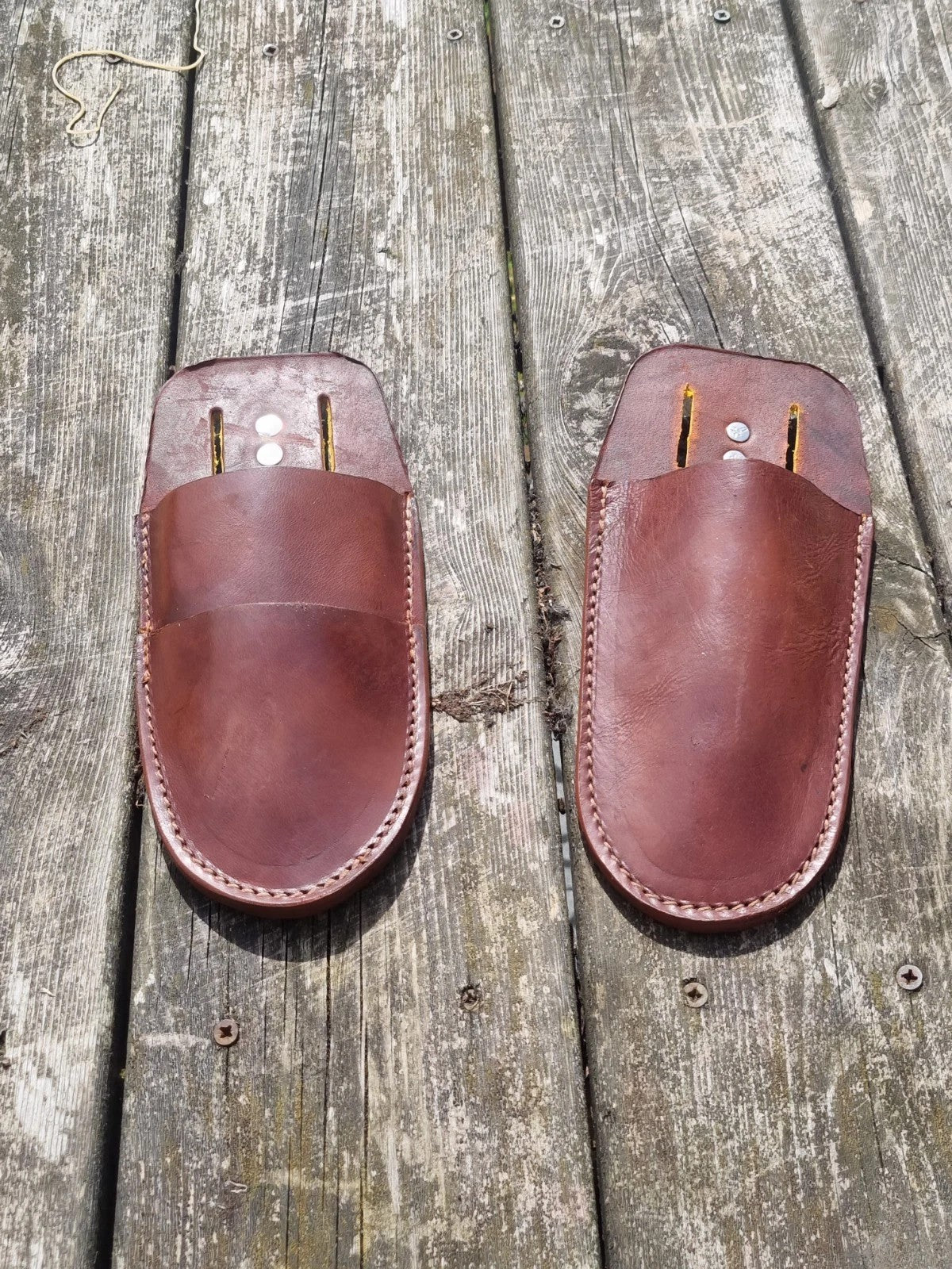 A pair of brown leather slip-on pouches or covers with stitched edges, laid flat on weathered wooden planks.