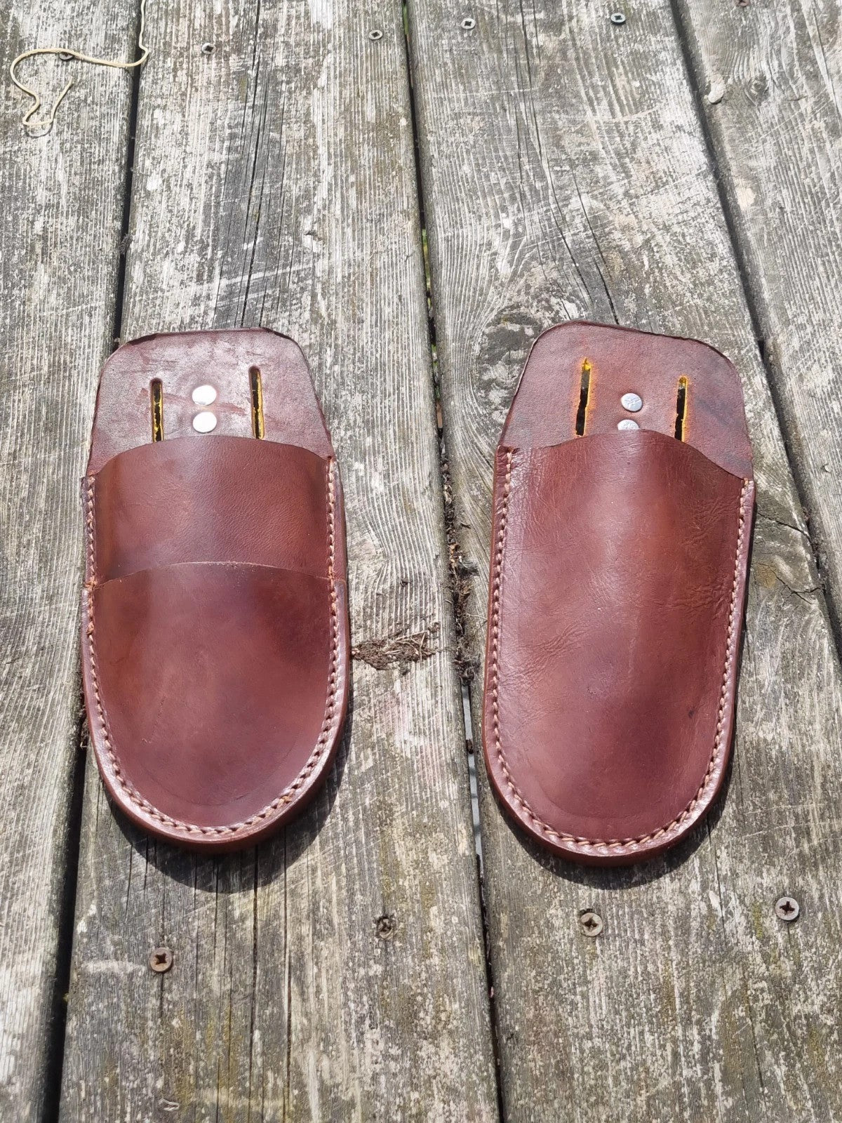 Two reddish-brown leather slip-on pouches or covers with visible stitching and metal rivets, placed on weathered wooden planks outdoors.