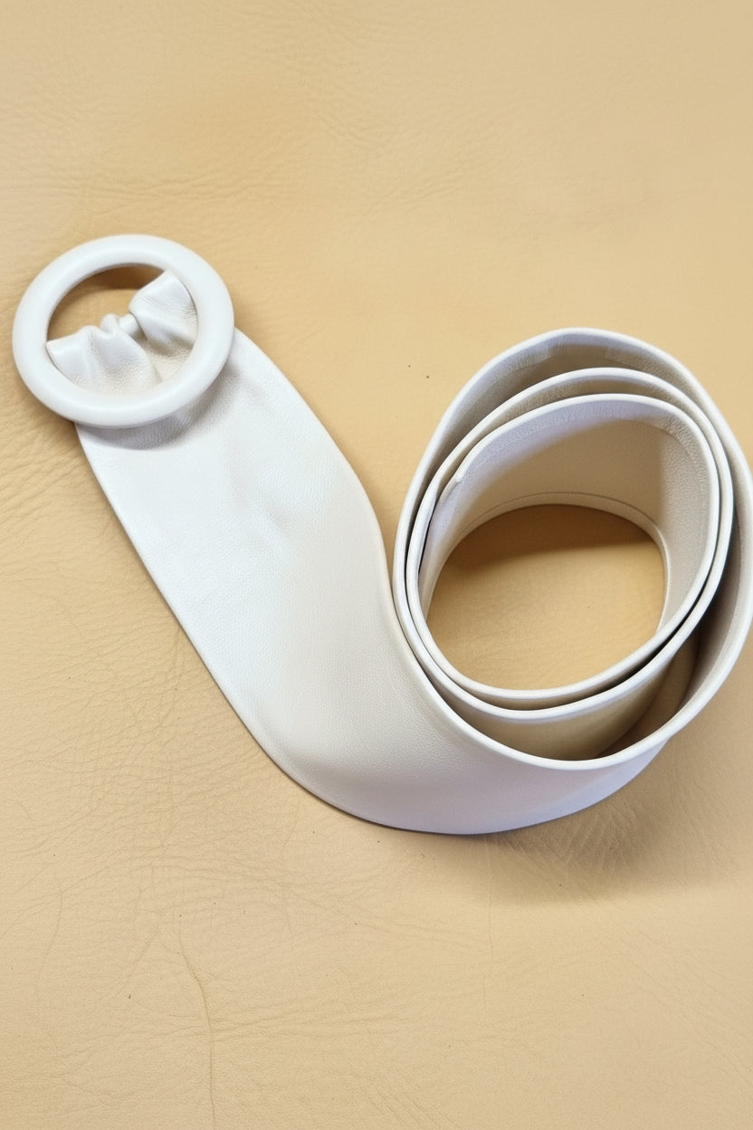 White women leather cinch belt with a wide strap and a large circular leather covered buckle, coiled on a tan textured leather background.