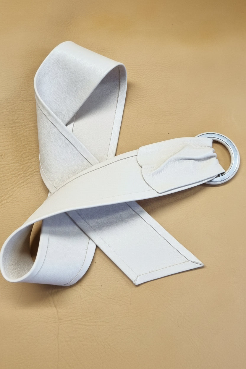 white leather cinch belt turned over to reveal finish, featuring a silver metal D-ring, arranged on a textured tan leather background.