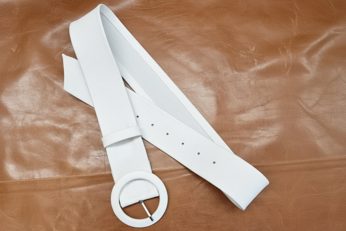 A white leather belt with a large circular ring buckle, laid out on a brown leather background.