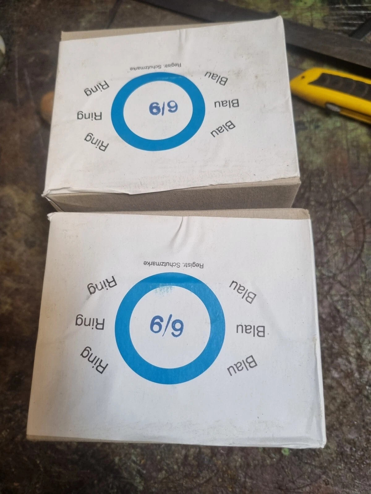 Two white paper boxes stacked on a dark textured surface, each printed with a blue ring graphic and the words '6/9' and 'Ring' repeated around it. A yellow utility knife is partially visible in the upper right corner.