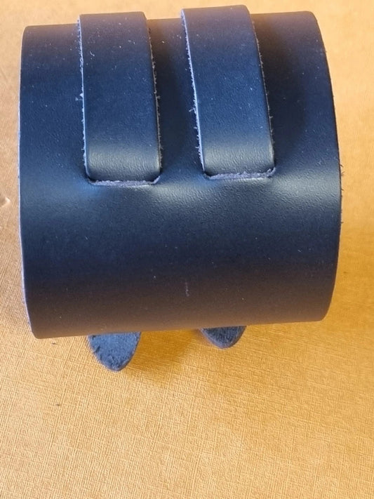 A black leather cuff bracelet with two raised tab details, displayed on a tan textured surface.