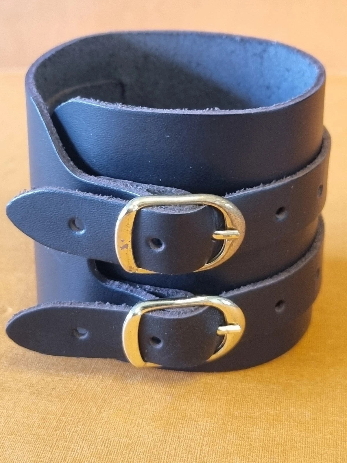 A black leather cuff bracelet with two adjustable straps and gold-tone metal buckles, displayed on a warm tan textured fabric background.