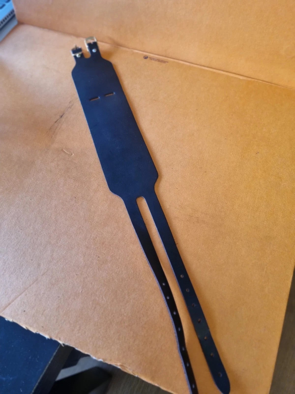 A black leather wrist cuff and support  resting flat and fully opened, photographed on a brown cardboard surface.