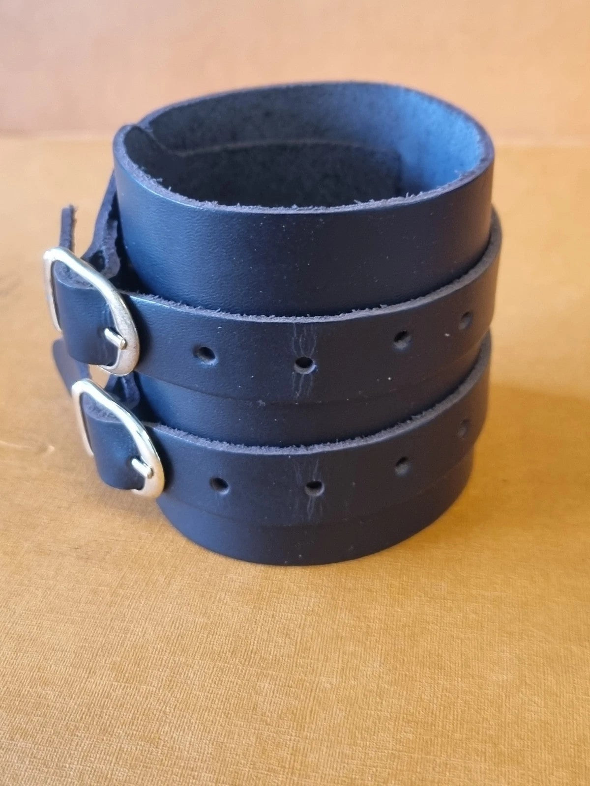 A black leather wrist cuff bracelet with two gold metal buckles, stacked on a tan textured fabric background.
