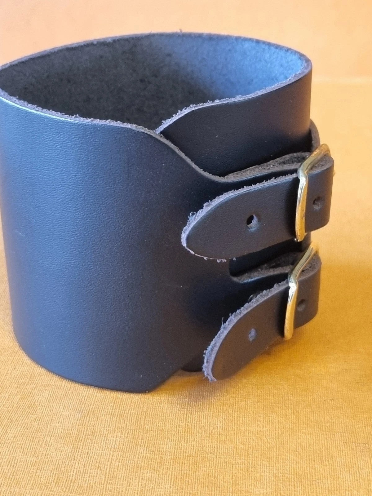 A wide black leather wrist cuff bracelet with two adjustable buckle straps, displayed on a textured tan fabric background.