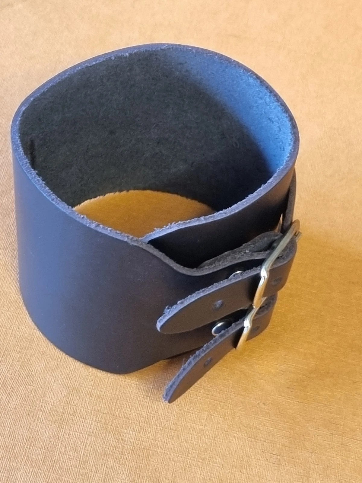 A wide black leather wrist cuff bracelet with a double-loop closure andgold metal buckles, photographed on a tan textured fabric background.