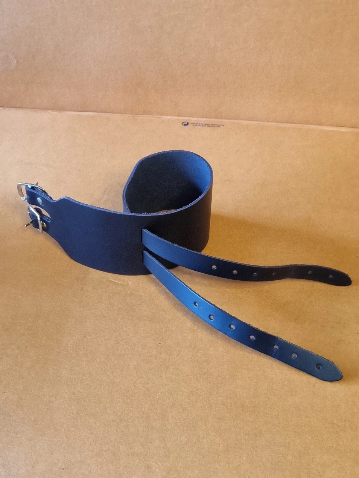 A black leather wrist cuff bracelet semi closed through adjustment holes, displayed on a brown cardboard background.